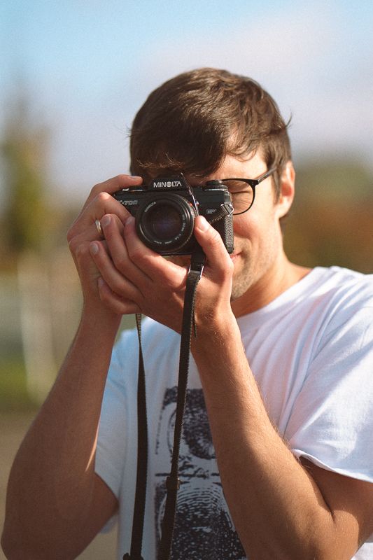 Young man in white t-shirt and glasses holding a Minolta film camera up to his eye outdoors.