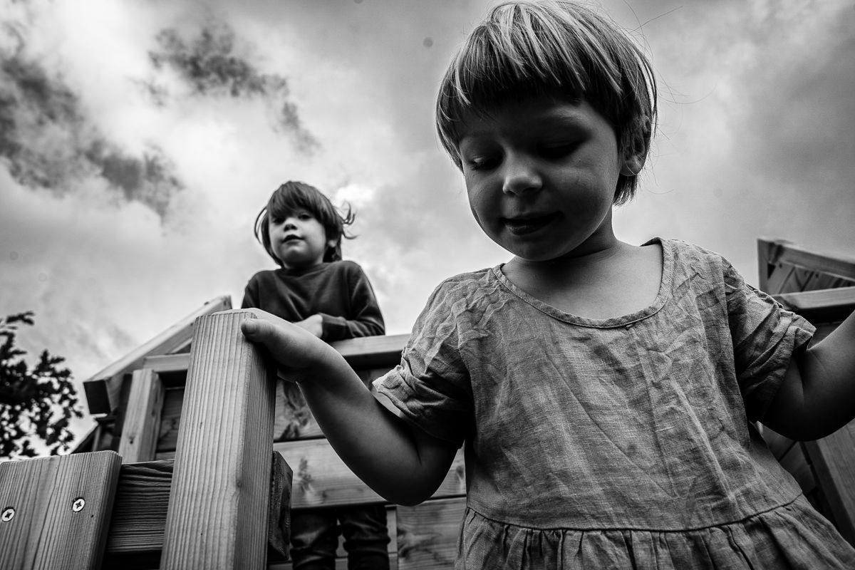 Black and white photo of two young children playing on a wooden playground structure under a cloudy sky.