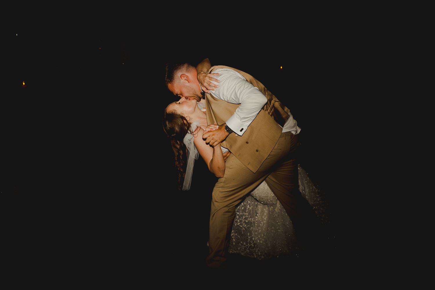 Couple sharing a romantic dip kiss during their wedding reception dance in a dimly lit outdoor setting.