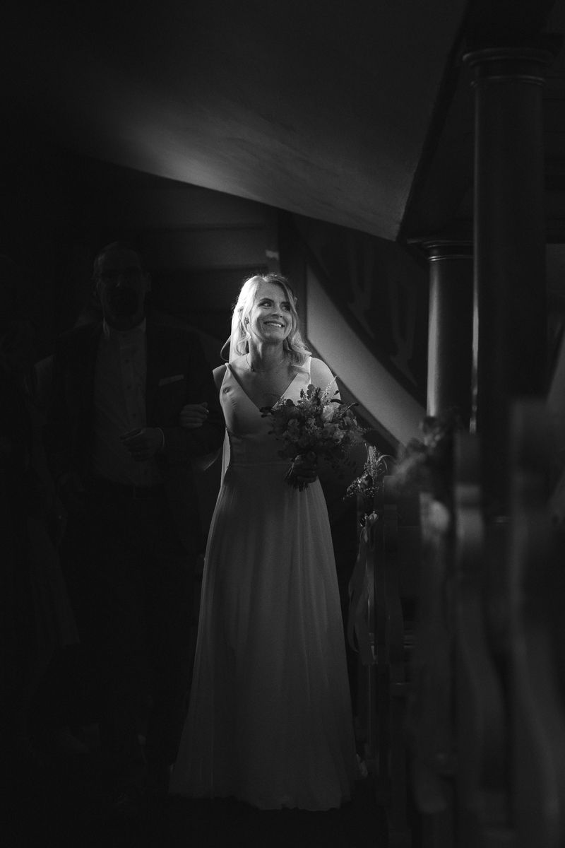 Black and white photo of a smiling bride in a white gown holding a bouquet, standing on a staircase in soft light.