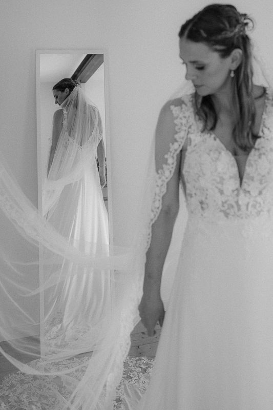 Bride in lace wedding gown with flowing veil admires her reflection in a full-length mirror, black and white photo.