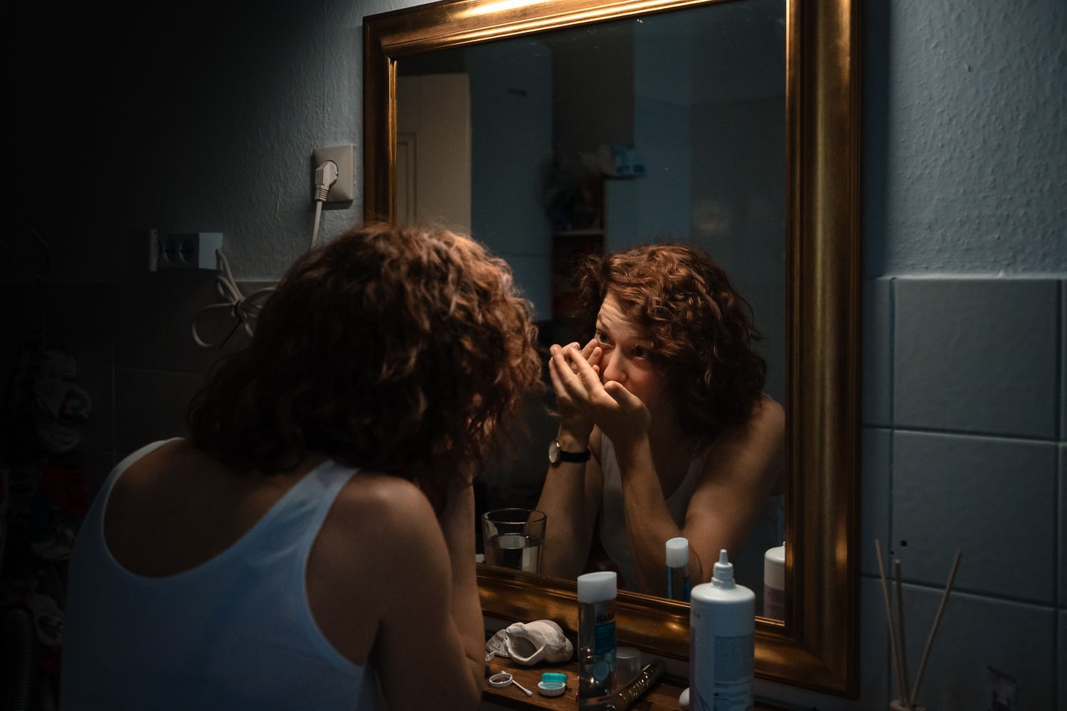 Person examining their face closely in a gold-framed bathroom mirror under dim lighting.