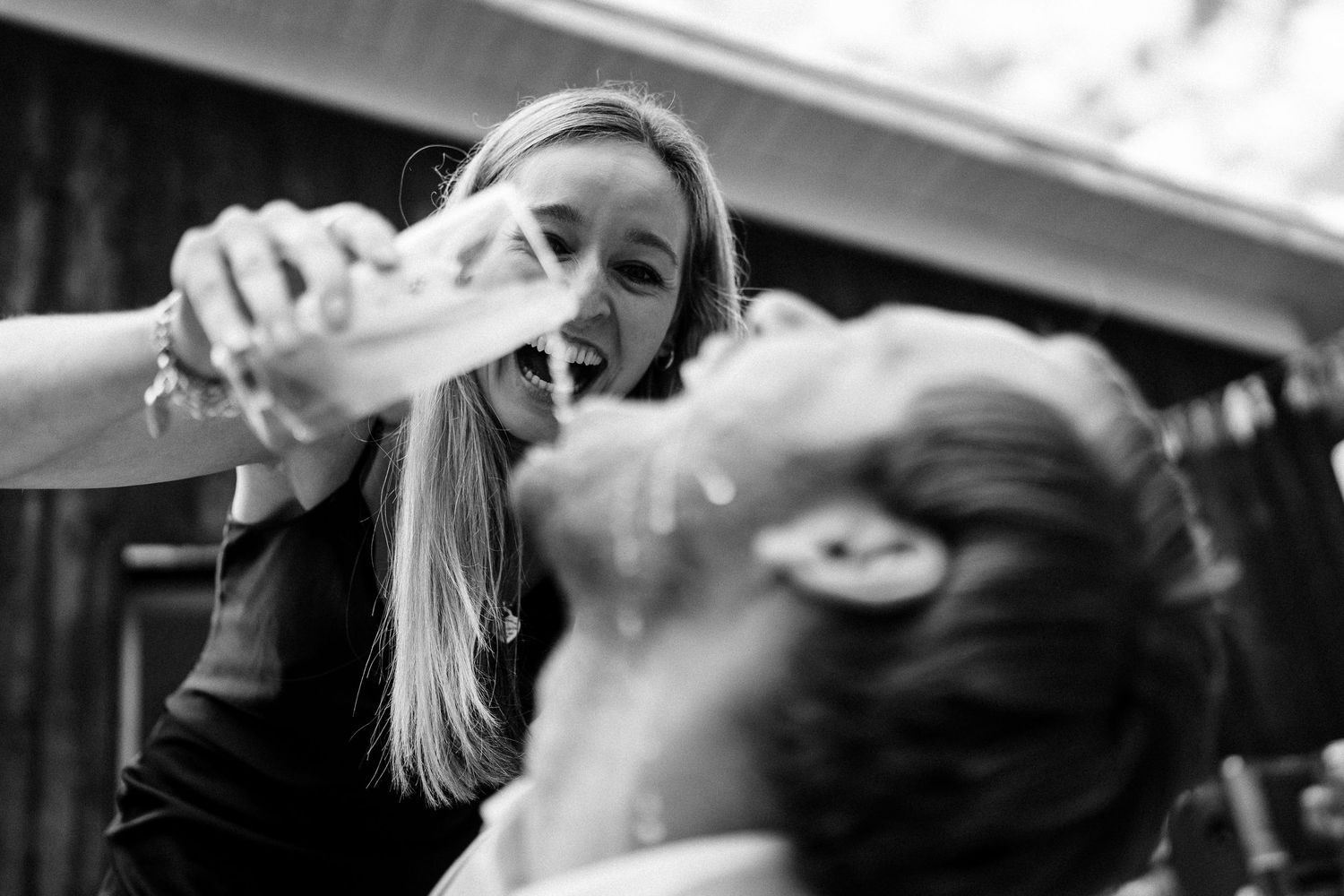 Woman playfully pouring drink into a laughing person's mouth at an outdoor event, black and white photo.