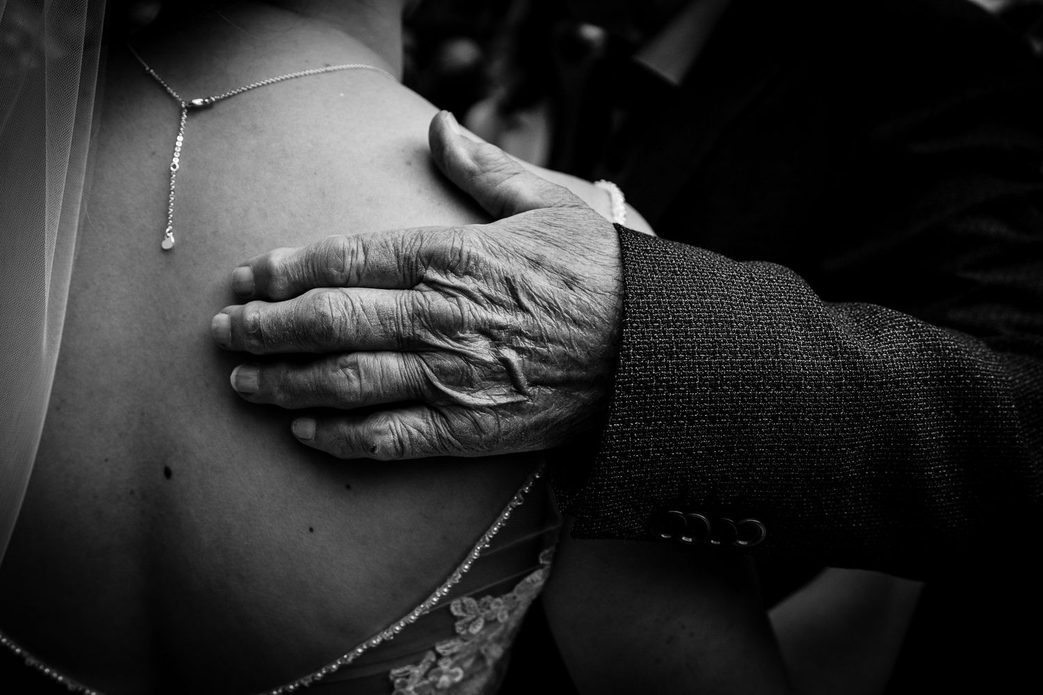 Black and white photo of an elderly hand resting on a young woman's bare shoulder, wearing a necklace.