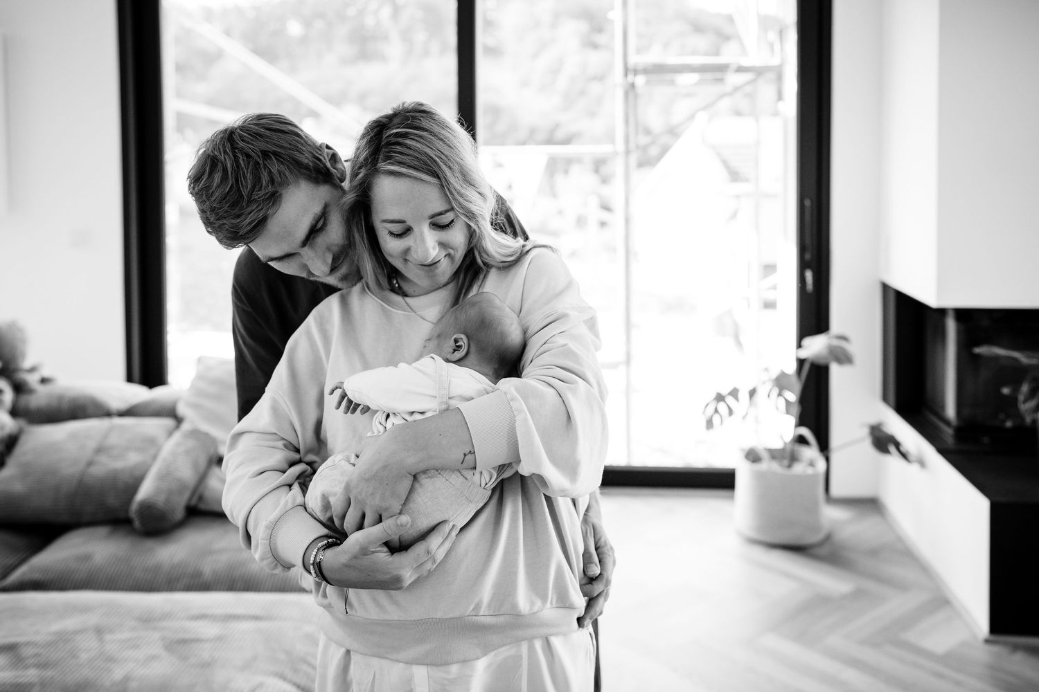 Schwarzweißfoto eines Paares, das zärtlich sein neugeborenes Baby in einem modernen Wohnzimmer in Freudenberg hält.
