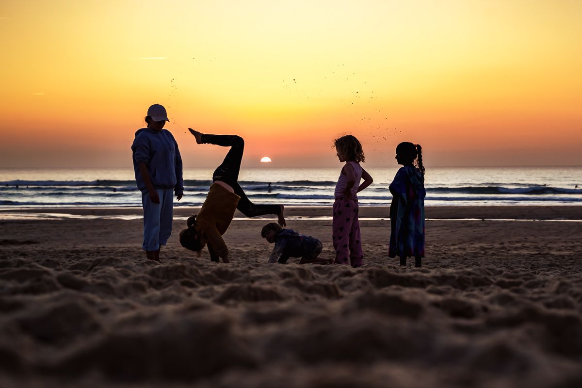 Silhouettes of family playing on sandy beach at golden sunset, child doing cartwheel while others watch near ocean waves.