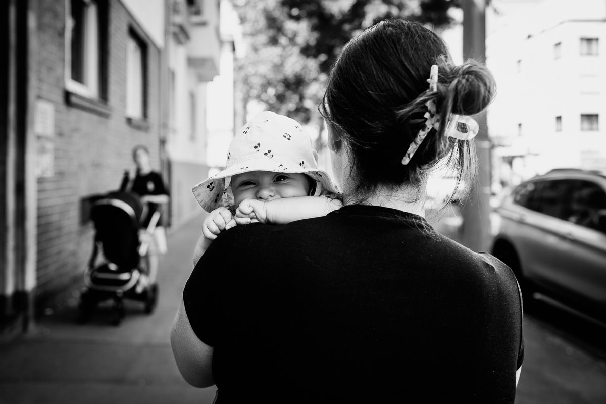 Black and white photo of a mother holding her baby in a hat outdoors on a city sidewalk.