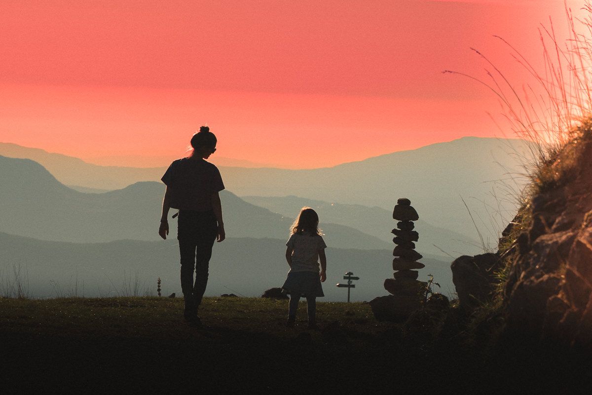 Silhouettes of a mother and child stand near a stone cairn at sunset with layered mountain ranges in the background.