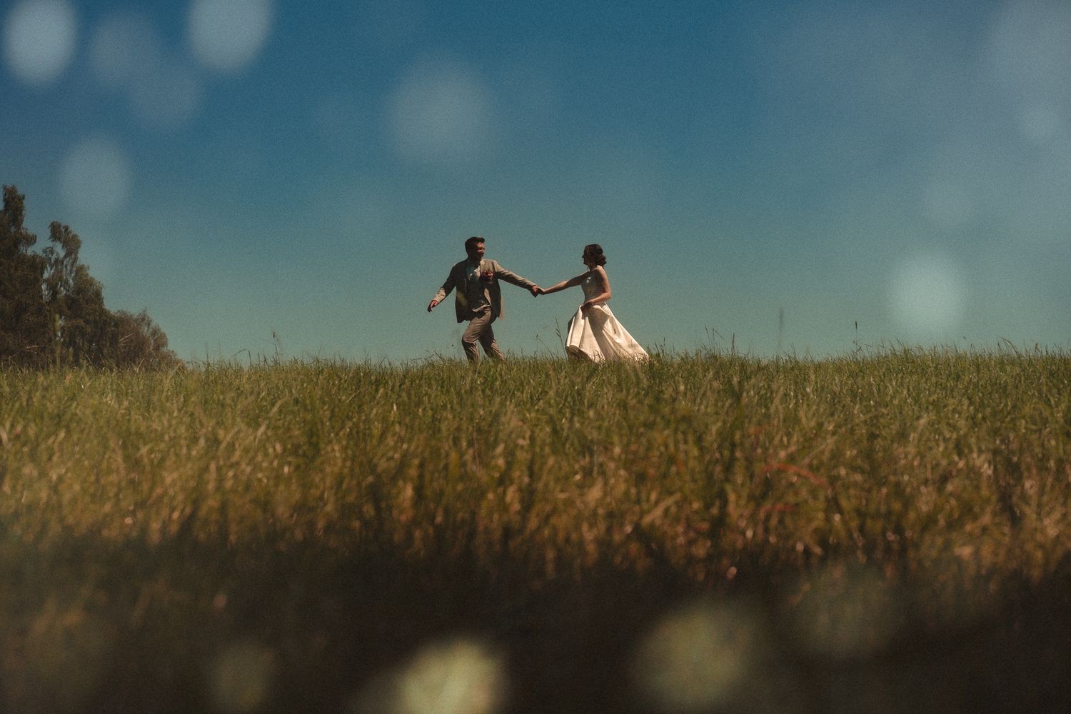 A couple holds hands and runs through a lush green field under a clear blue sky on a sunny day.