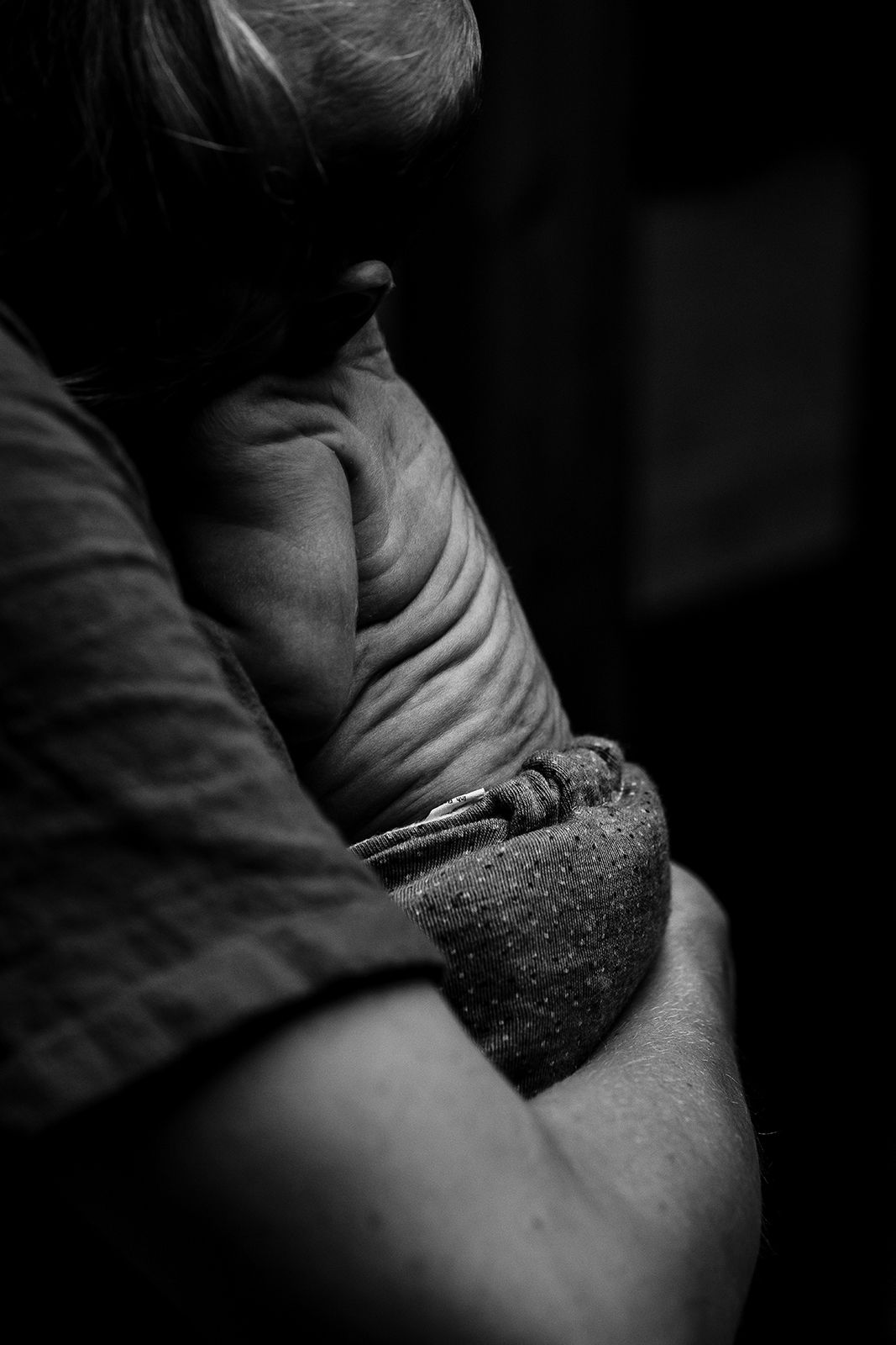 Black and white close-up of an elderly person with arms crossed, showing aged skin texture in dramatic lighting.