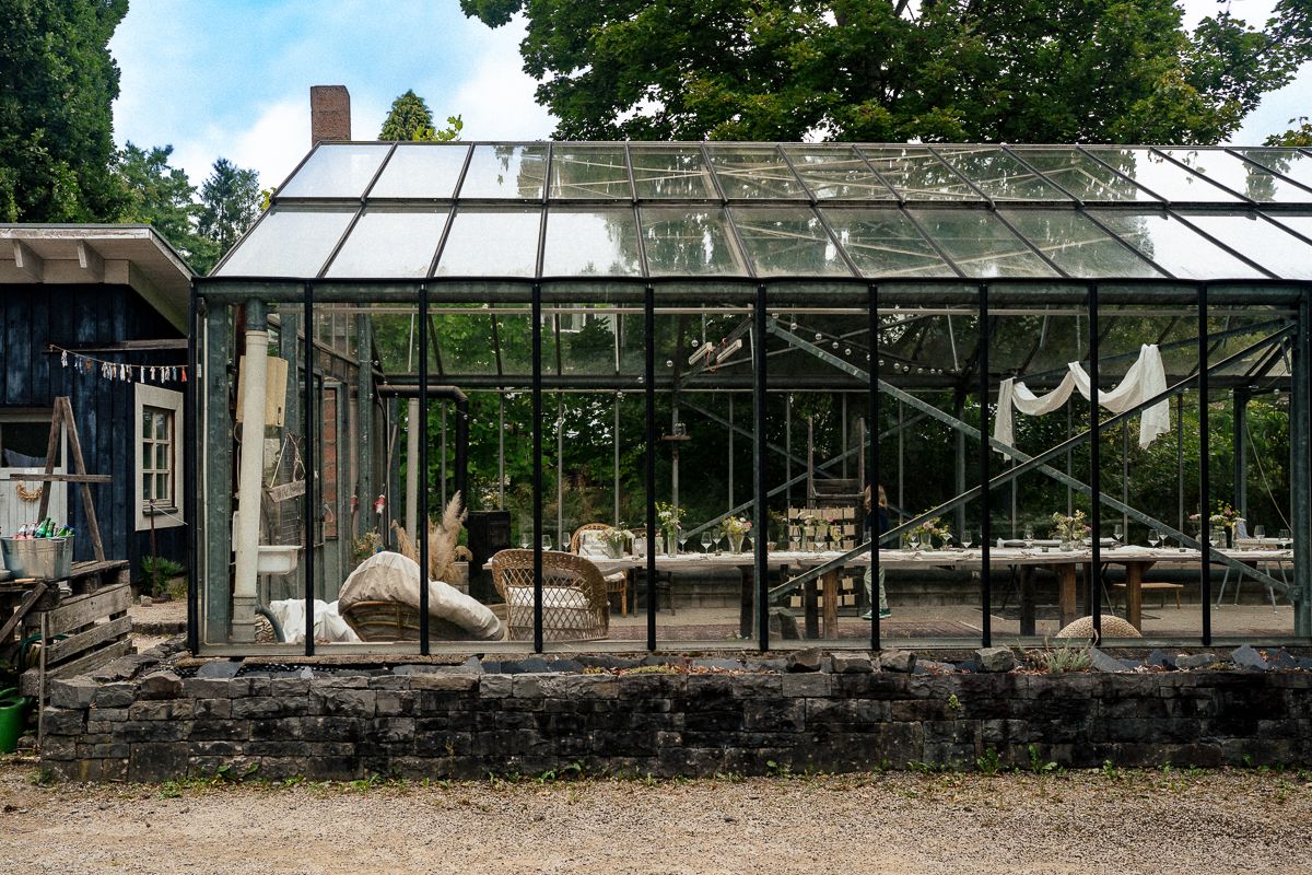 Vintage black metal greenhouse with glass panels, rustic stone foundation, and elegant interior furniture in a garden setting.