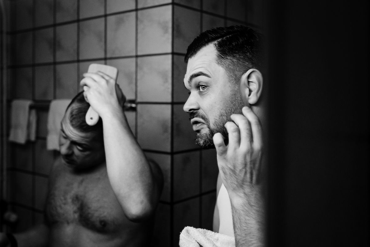 Black and white photo of a man examining his beard in a bathroom mirror, touching his face thoughtfully.
