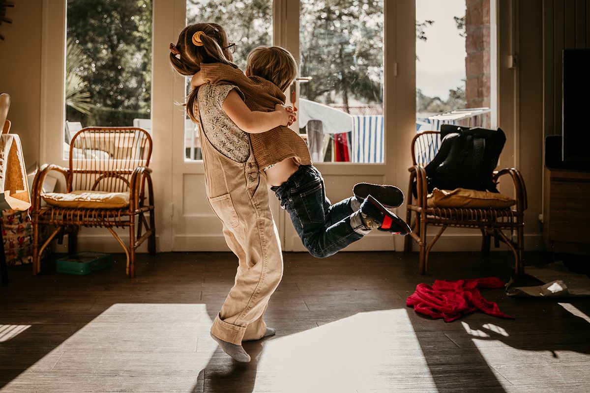A joyful child being lifted and hugged by an adult in a sunlit living room with rattan chairs.