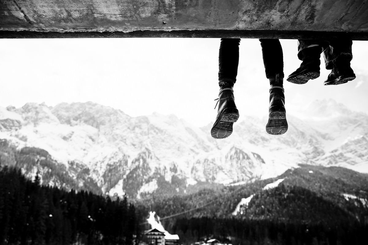 Two people sit on a ledge with legs dangling over a snowy mountain landscape in black and white.