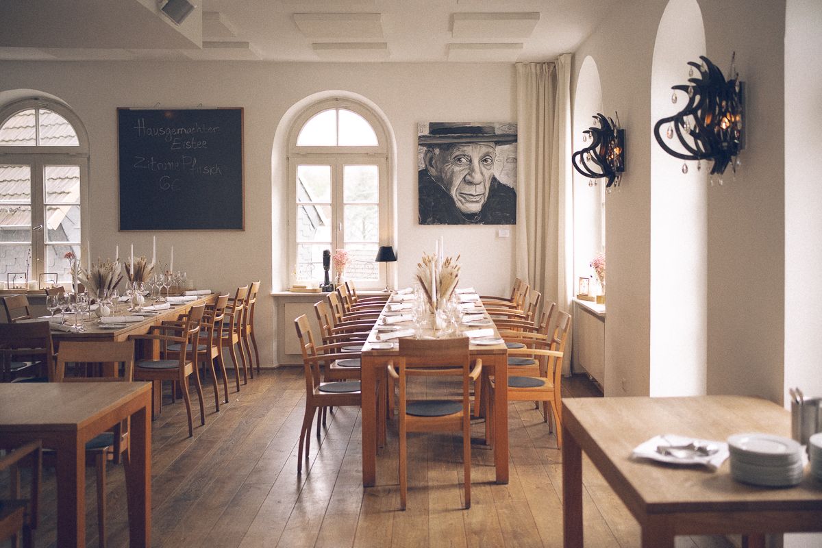 Elegant restaurant dining room with wooden tables, chairs, arched windows, and a portrait painting on white walls.