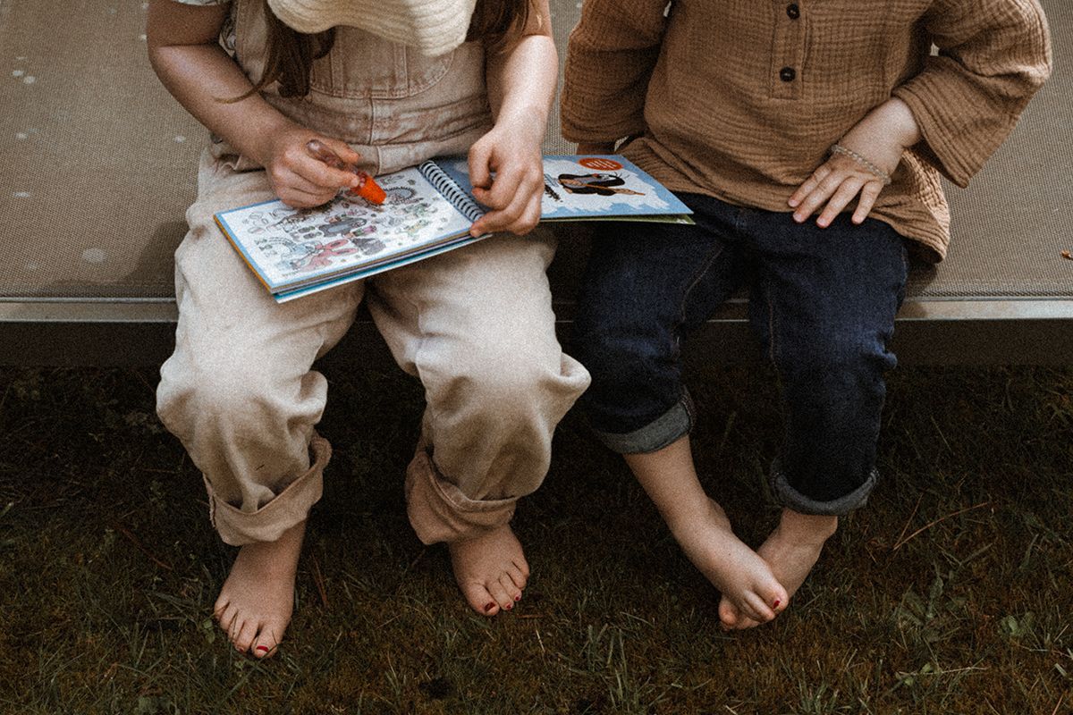 Two barefoot children sitting side by side reading a colorful book together outdoors.