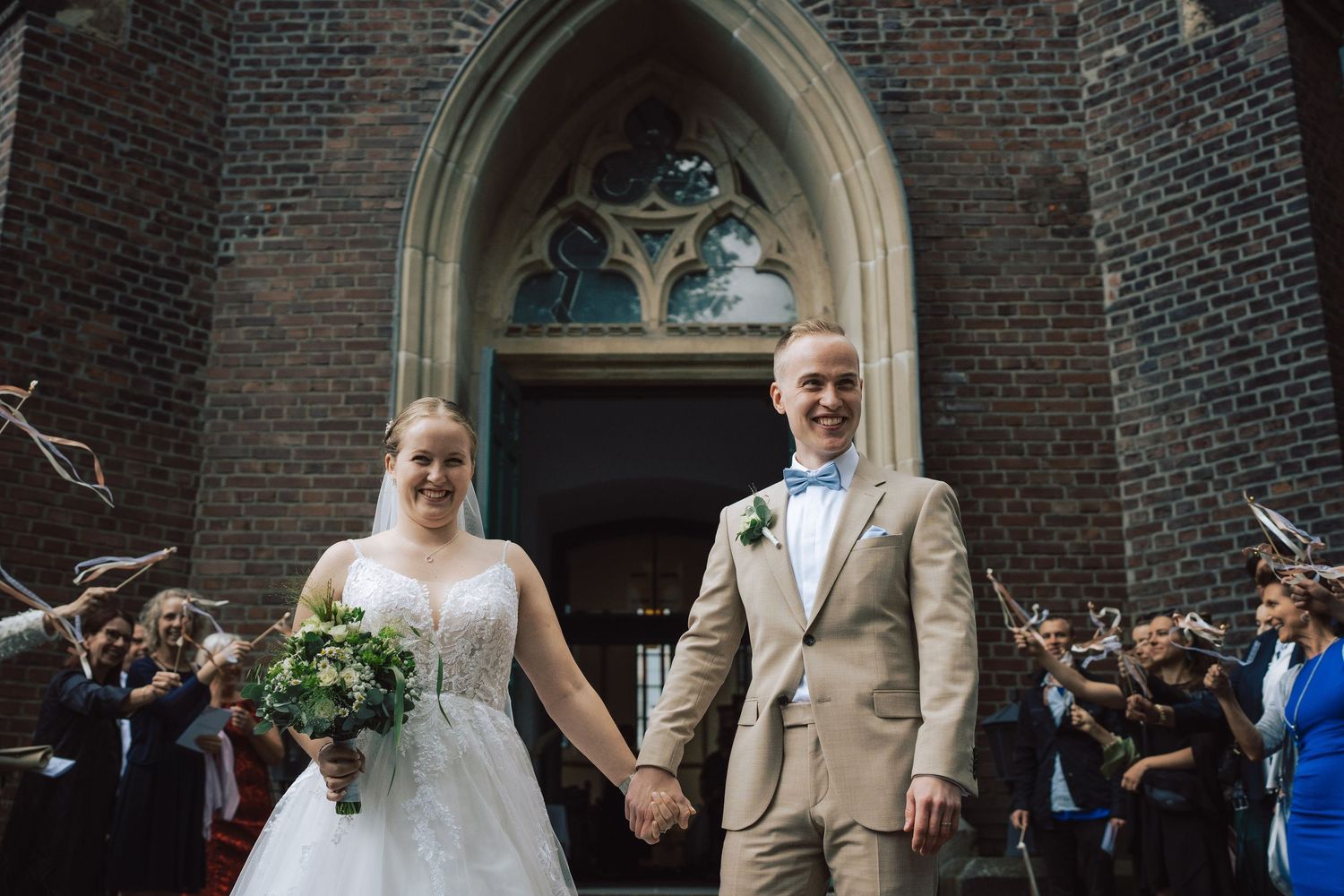 Newlywed couple holding hands outside a brick church, bride in white gown with bouquet, groom in beige suit with blue bow tie.