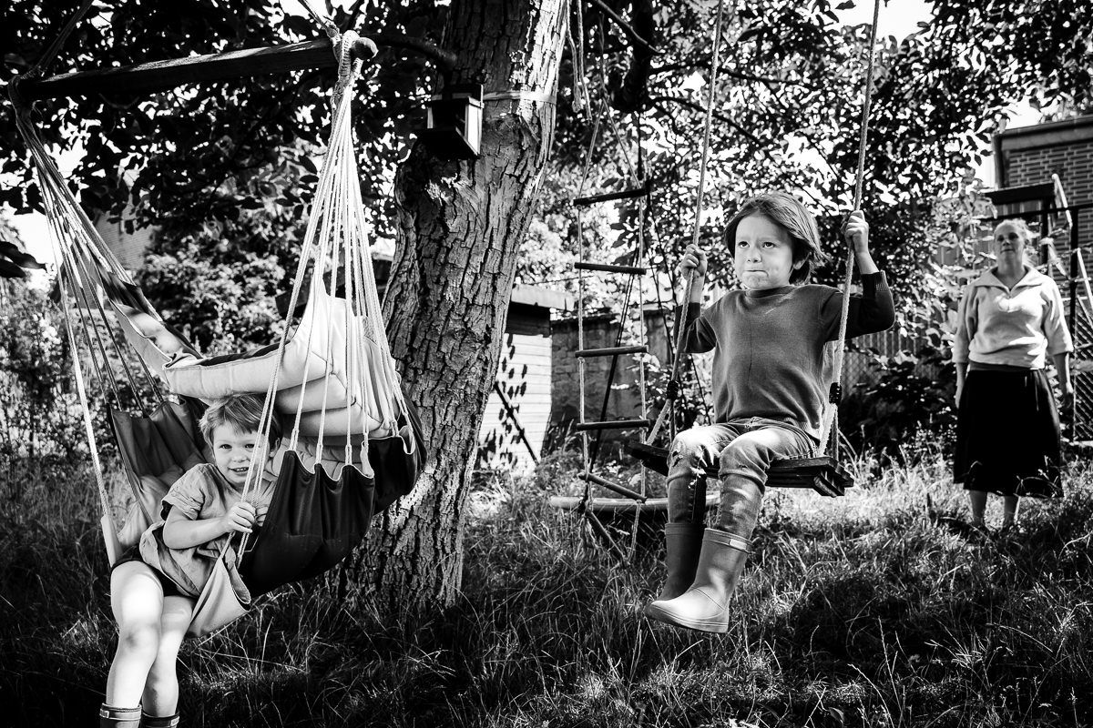 Black and white photo of two children on swings in a garden with a woman standing nearby.