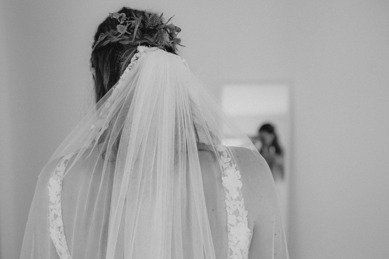Black and white photo of a bride in a lace wedding dress and veil, viewed from behind, standing before a mirror.