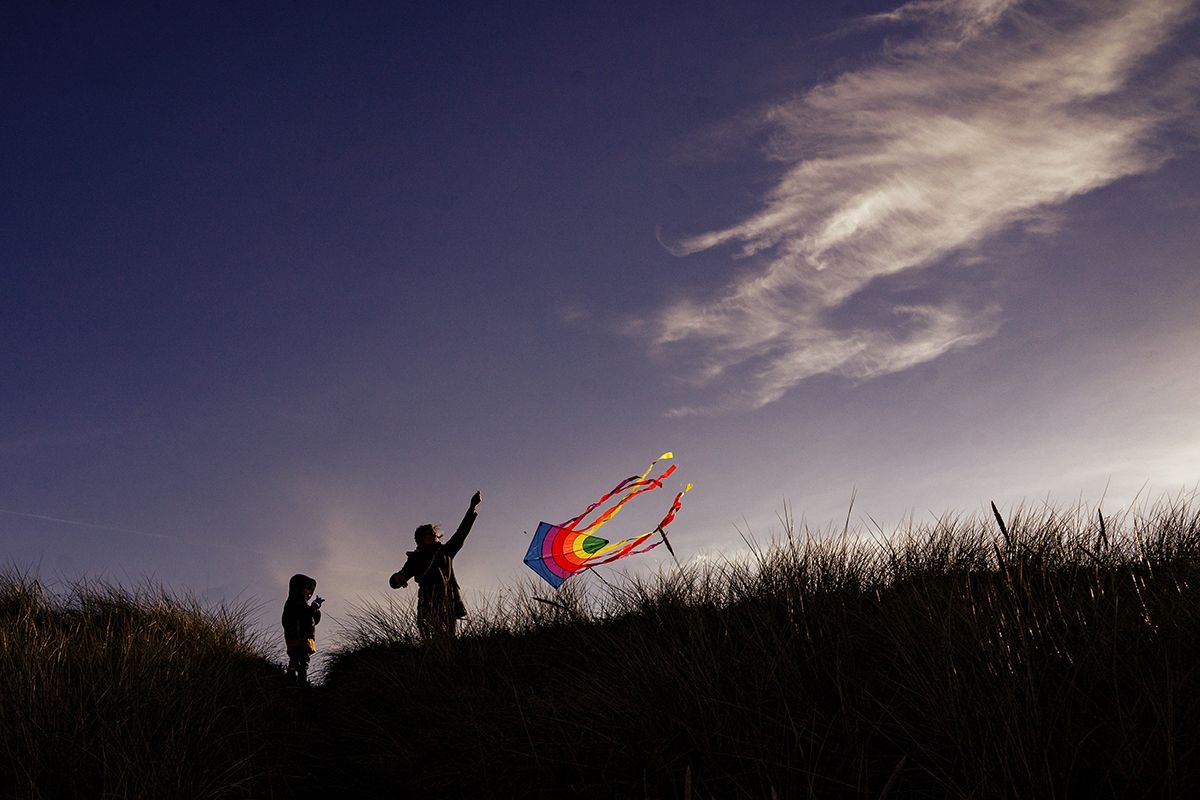 Two silhouetted figures fly a colorful rainbow kite against a dramatic twilight sky in an open grassy field.