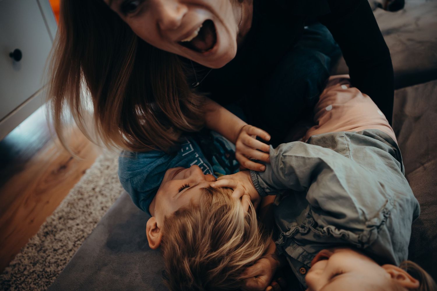 A laughing mother playfully tickles her two young children on the floor at home.
