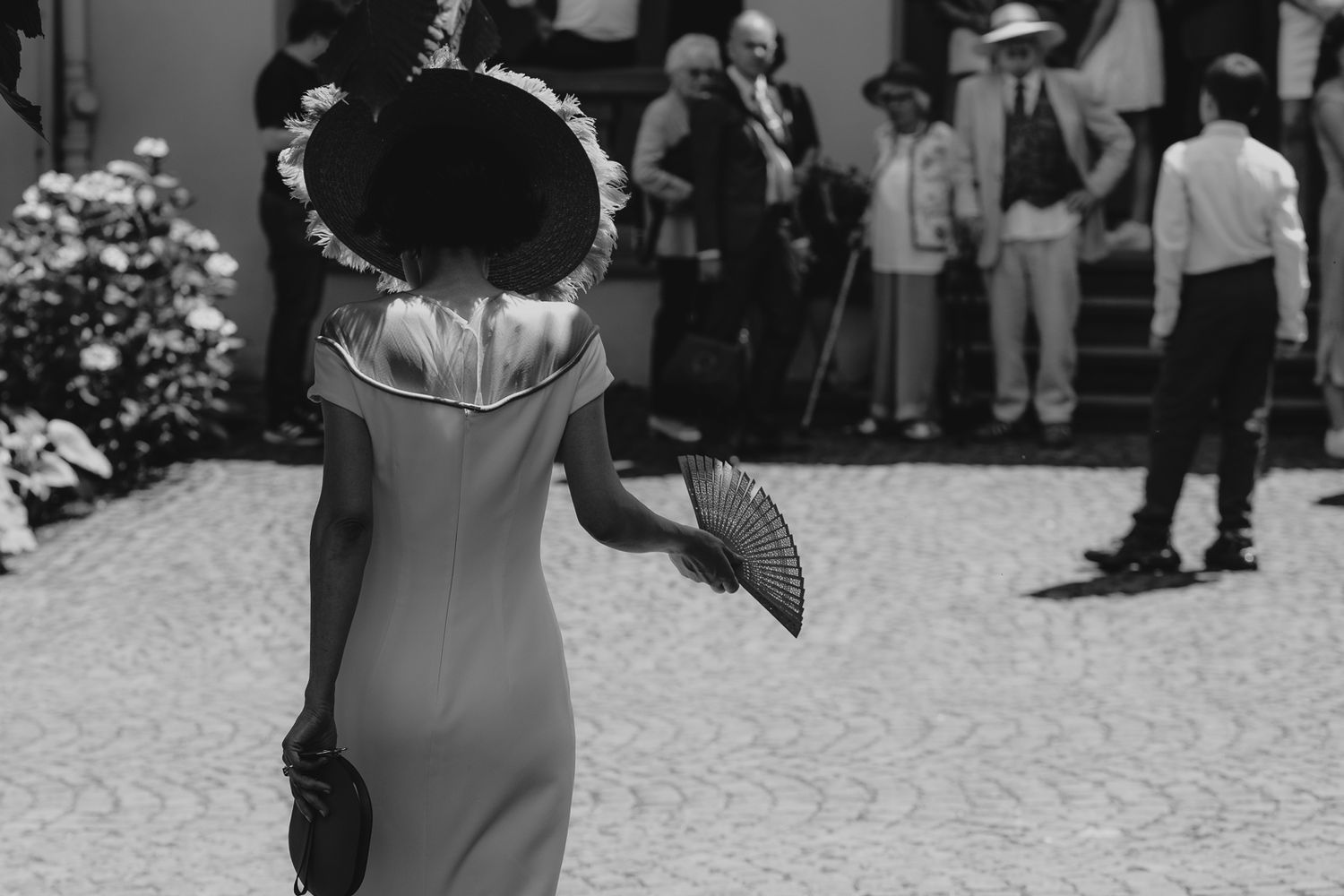 Elegant woman in vintage dress and wide-brim hat holding a fan at an outdoor gathering, black and white photo.