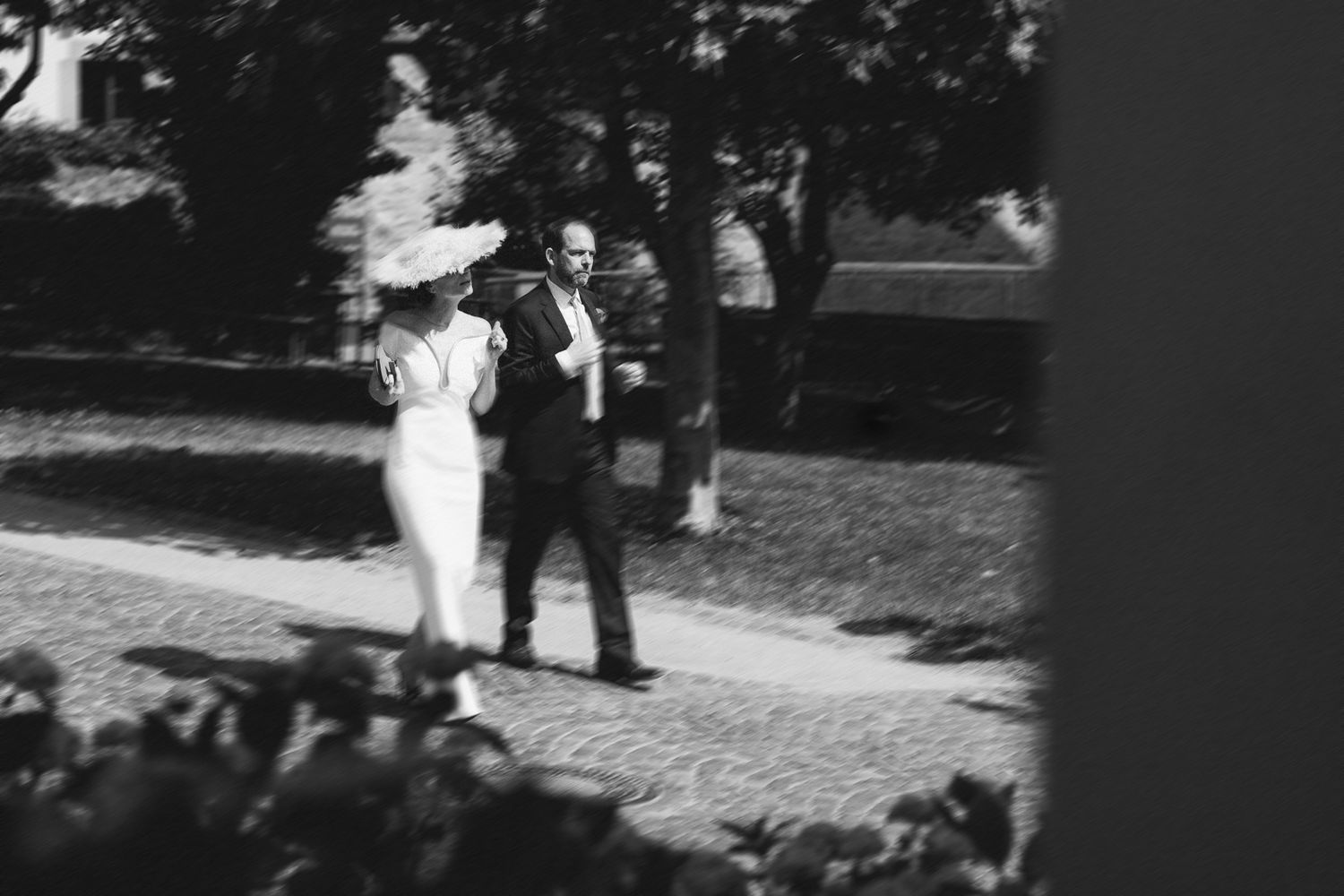 Black and white photo of a bride and groom walking outdoors on a cobblestone path surrounded by trees.