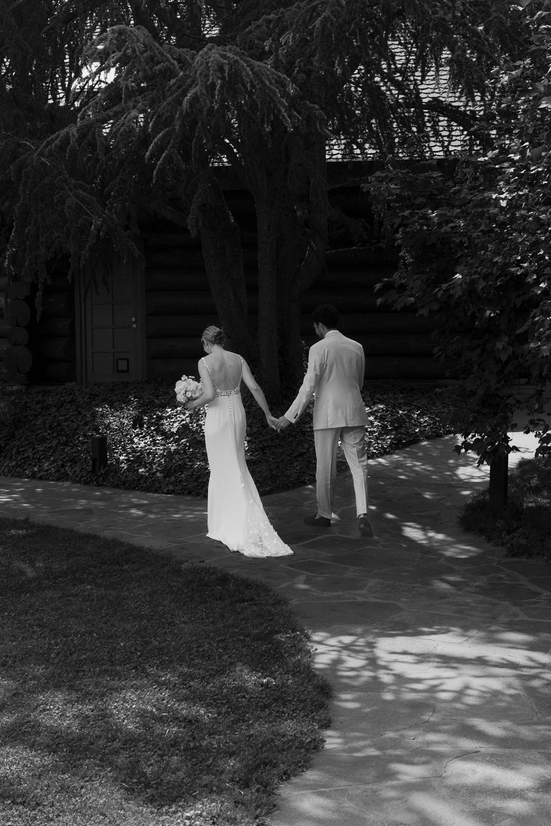 Black and white photo of bride and groom holding hands walking toward a rustic building surrounded by lush trees.