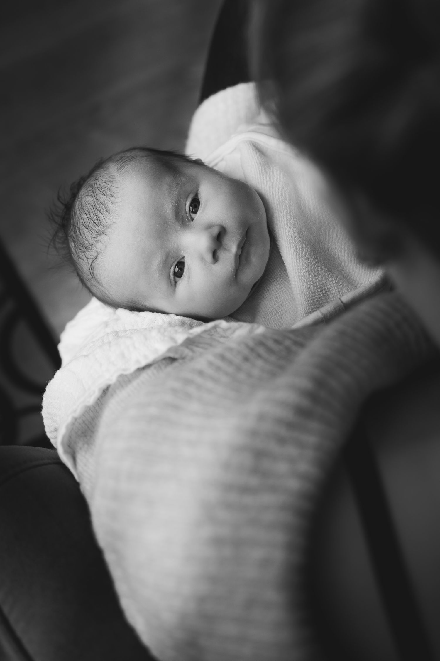 Schwarzweißfoto eines neugeborenen Babys, eingewickelt in eine weiße Decke, mit wachen Augen nach oben blickend.