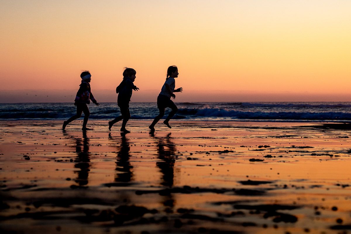 Three children running along a beach at sunset, their silhouettes reflected on wet sand with golden orange sky.