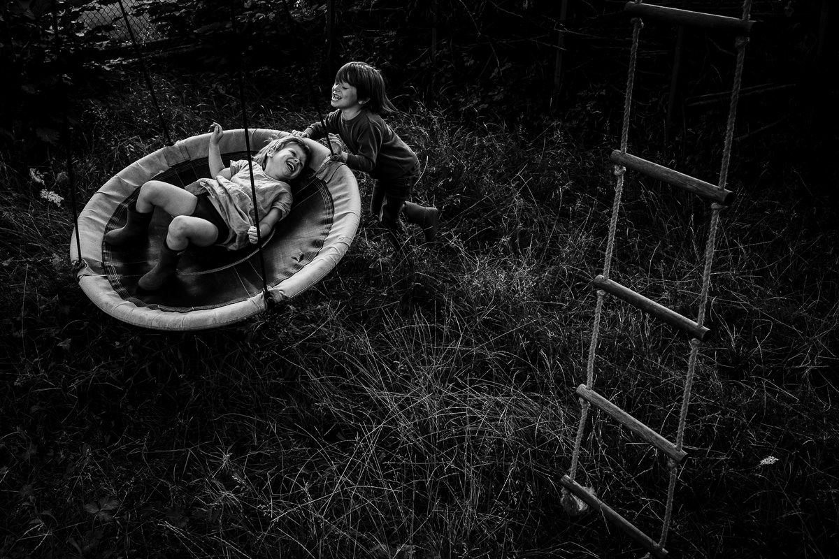 Schwarzweißfoto zweier Kinder beim Spielen im Freien, eines sitzt in einer runden Schaukel, während das andere daneben steht.