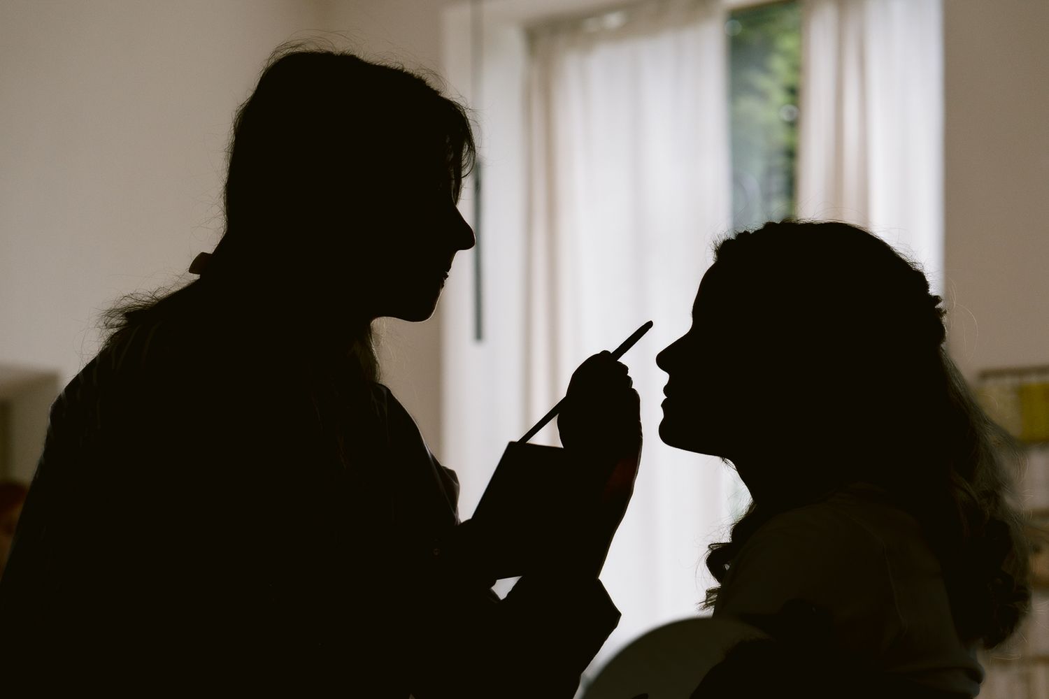 Silhouette of makeup artist applying cosmetics to a client against a bright window backdrop.