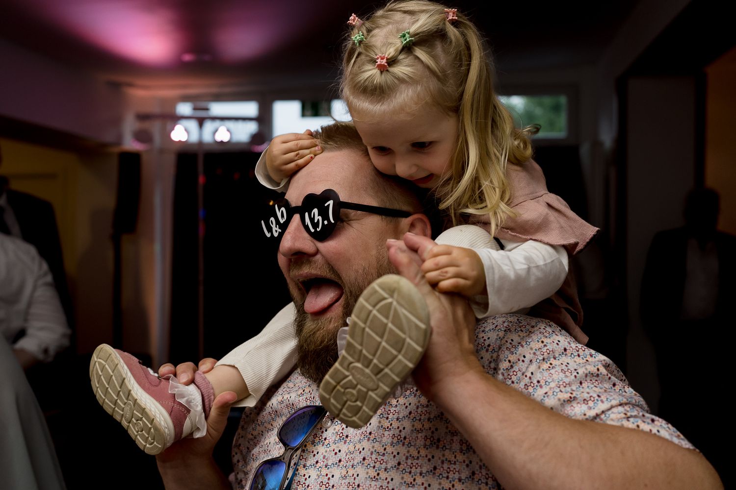 Man with heart-shaped sunglasses laughing as a little blonde girl sits on his shoulders at a wedding reception.