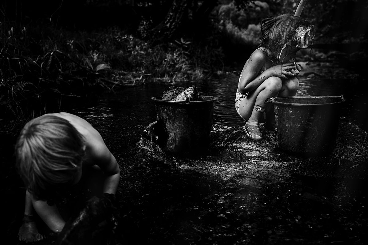 Black and white photo of two children playing outdoors near metal buckets in a natural woodland setting.