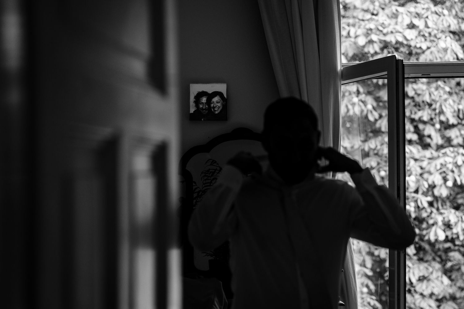 Black and white photo of a person adjusting their tie near a window with ivy, with a framed photo visible on the wall.