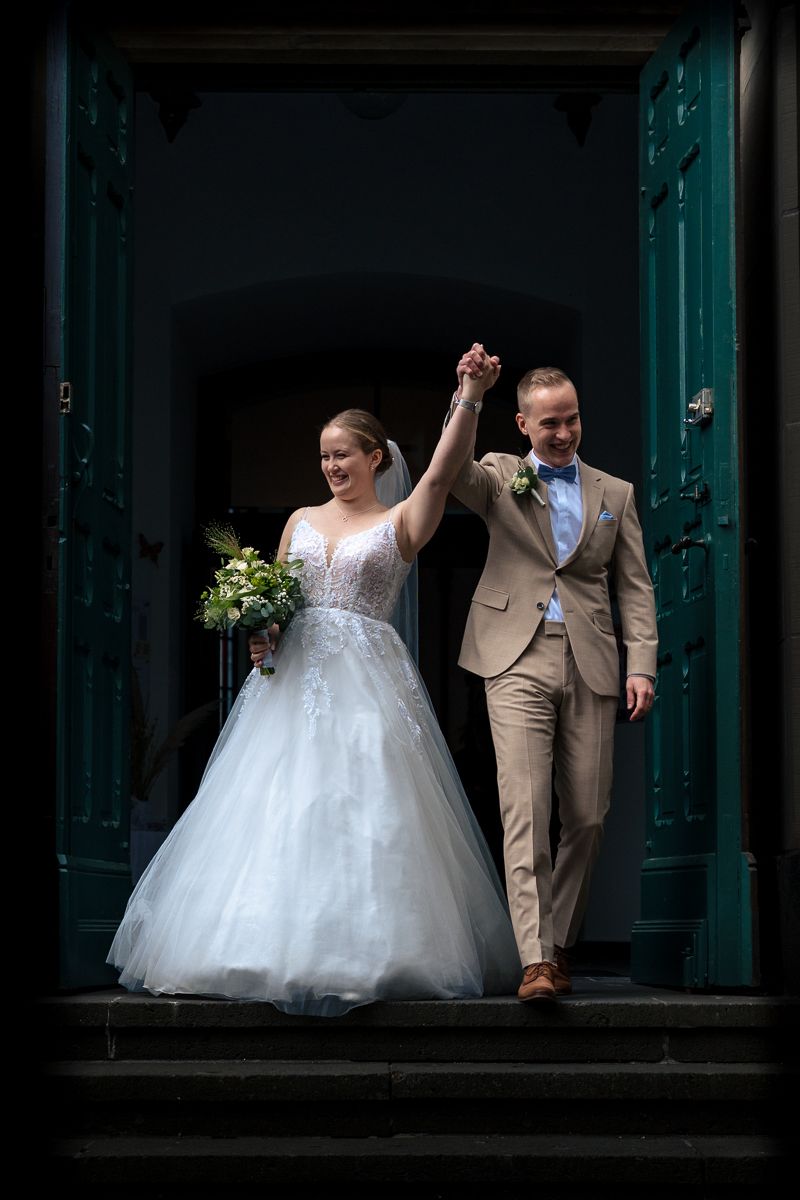 Bride and groom holding hands raised, exiting church doors, bride in white gown holding bouquet, groom in tan suit.