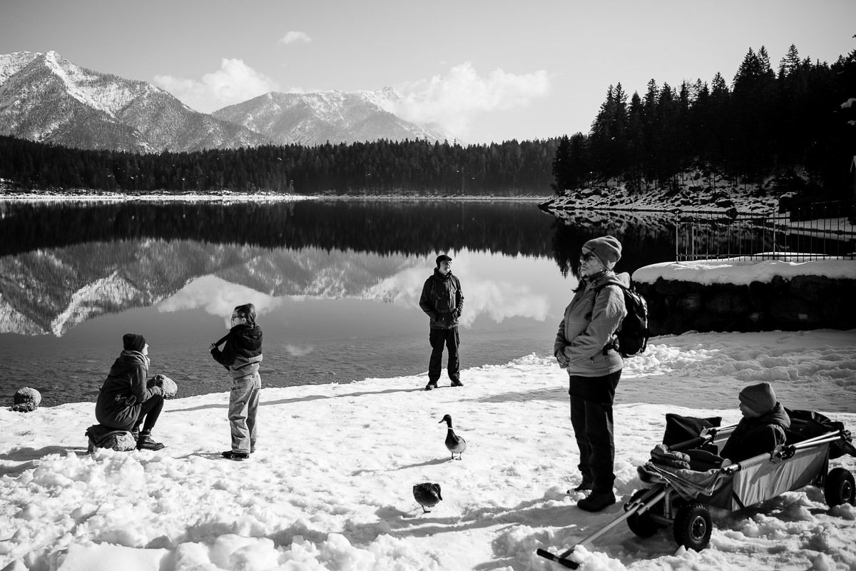 Group of people standing on snowy shore beside calm mountain lake with forest and peaks reflected in water.