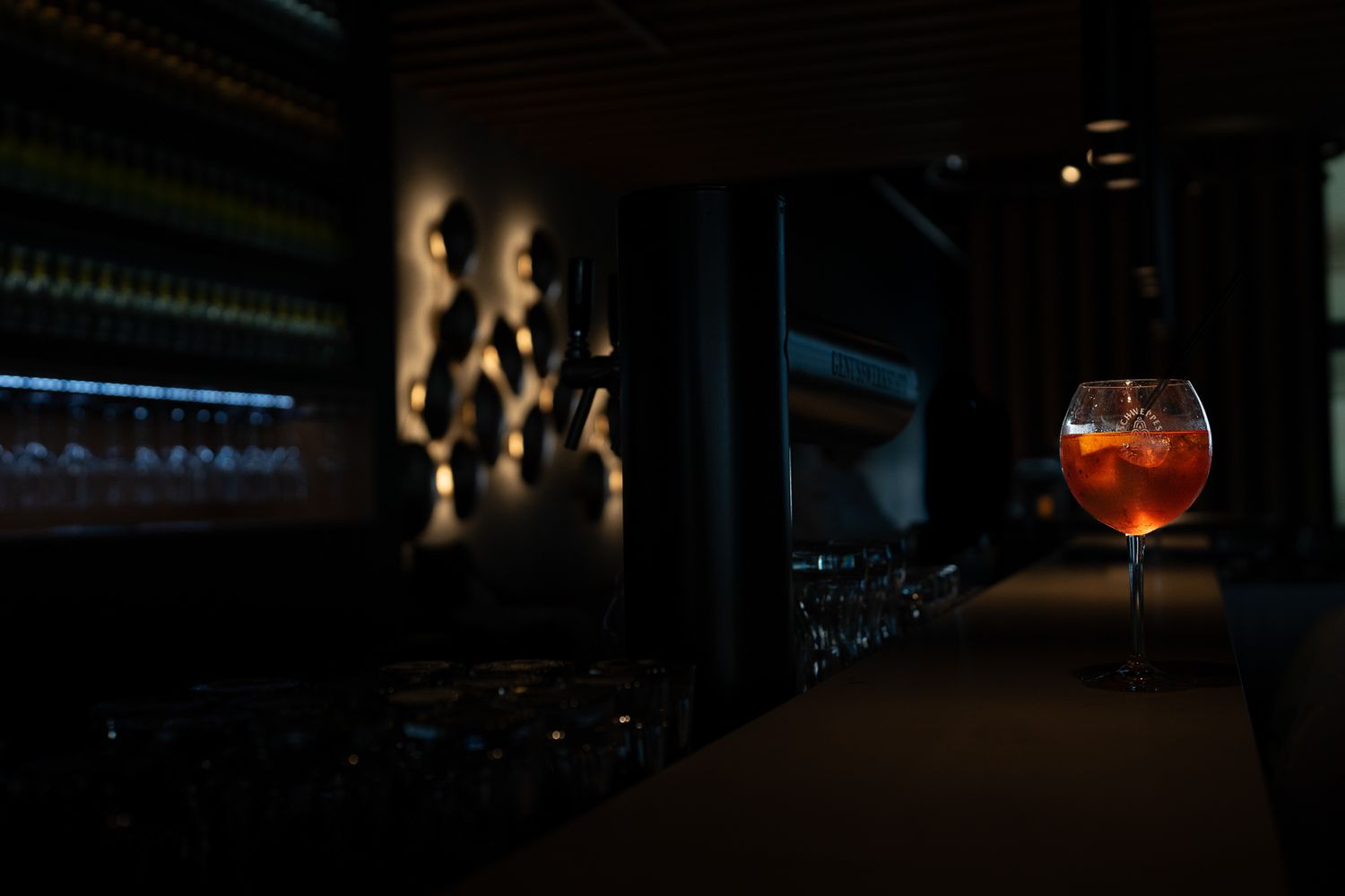 A glowing orange cocktail in a large wine glass sits on a dark bar counter in a dimly lit upscale lounge.