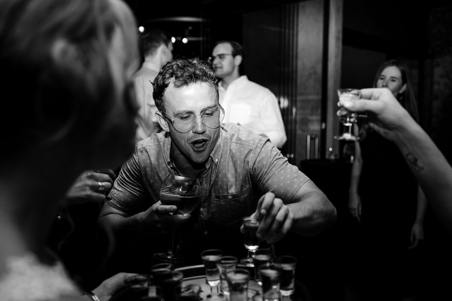 Black and white photo of a man holding a drink at a bar, surrounded by friends toasting with glasses.
