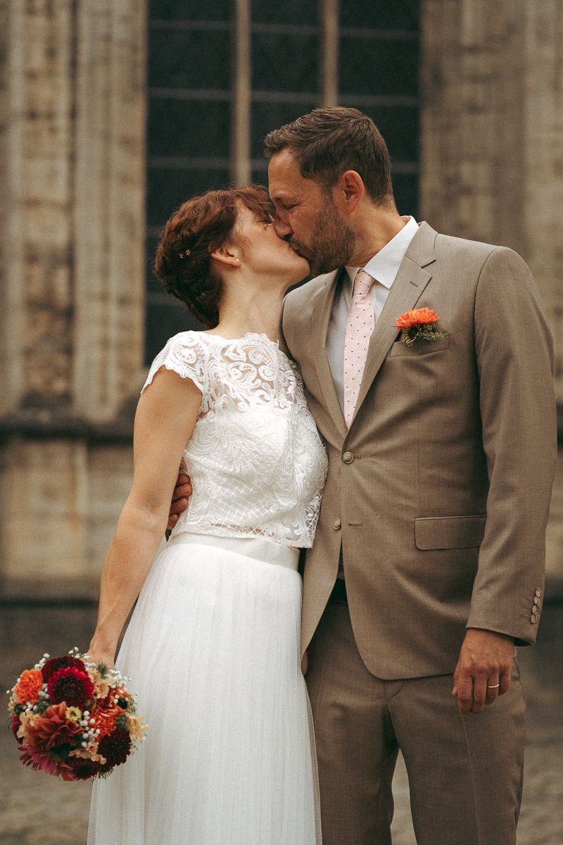 Braut im Spitzenhochzeitskleid und Bräutigam im beigefarbenen Anzug küssen sich vor der Kirche in Braunschweig.