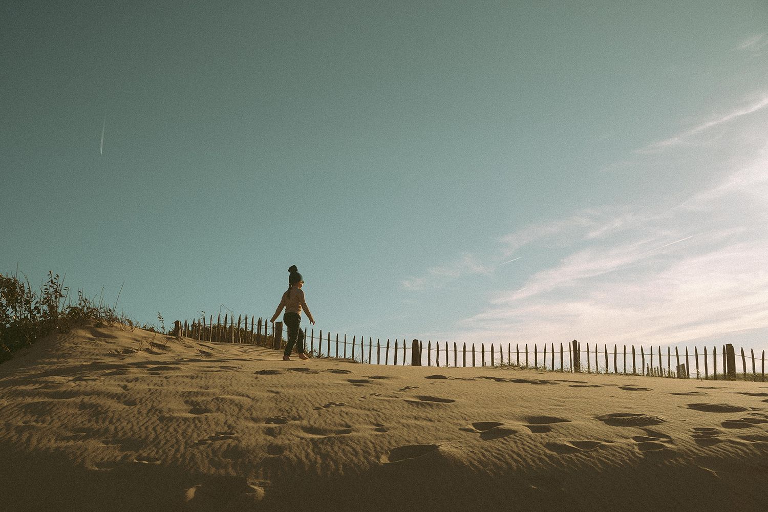 A lone person walks along a sandy dune beside a wooden fence under a blue sky.