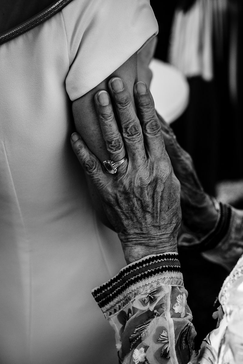 Black and white photo of an elderly hand with rings and bracelets resting gently on a bride's shoulder.