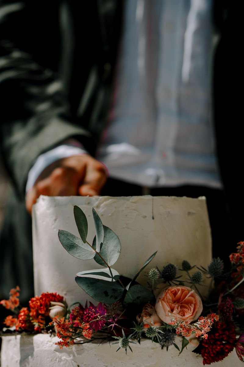 Elegant white wedding cake adorned with fresh roses, thistles, and eucalyptus leaves, with a groom in a suit standing behind.