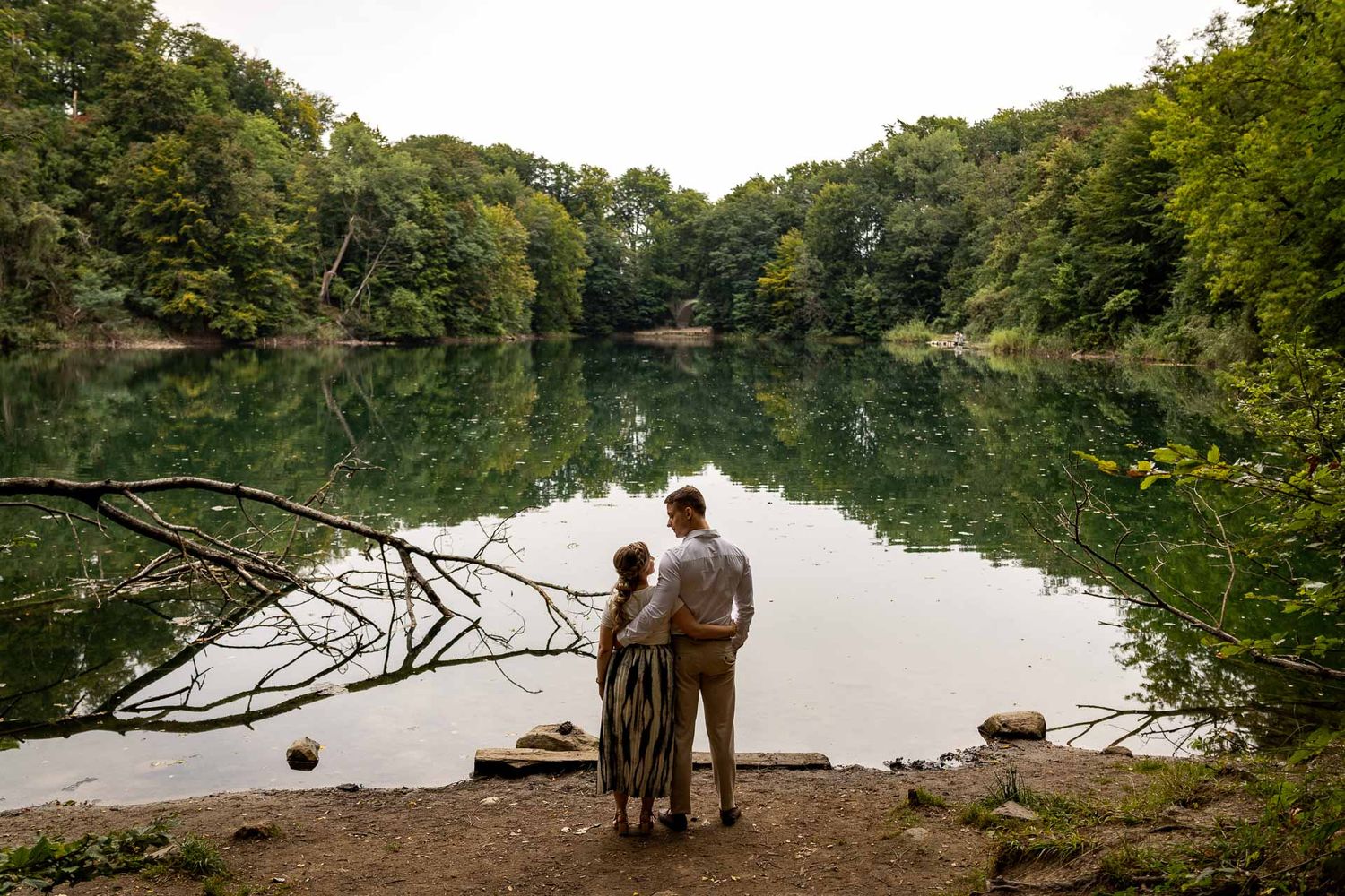 Panoramic view of a serene lake with trees and people standing on the shore.