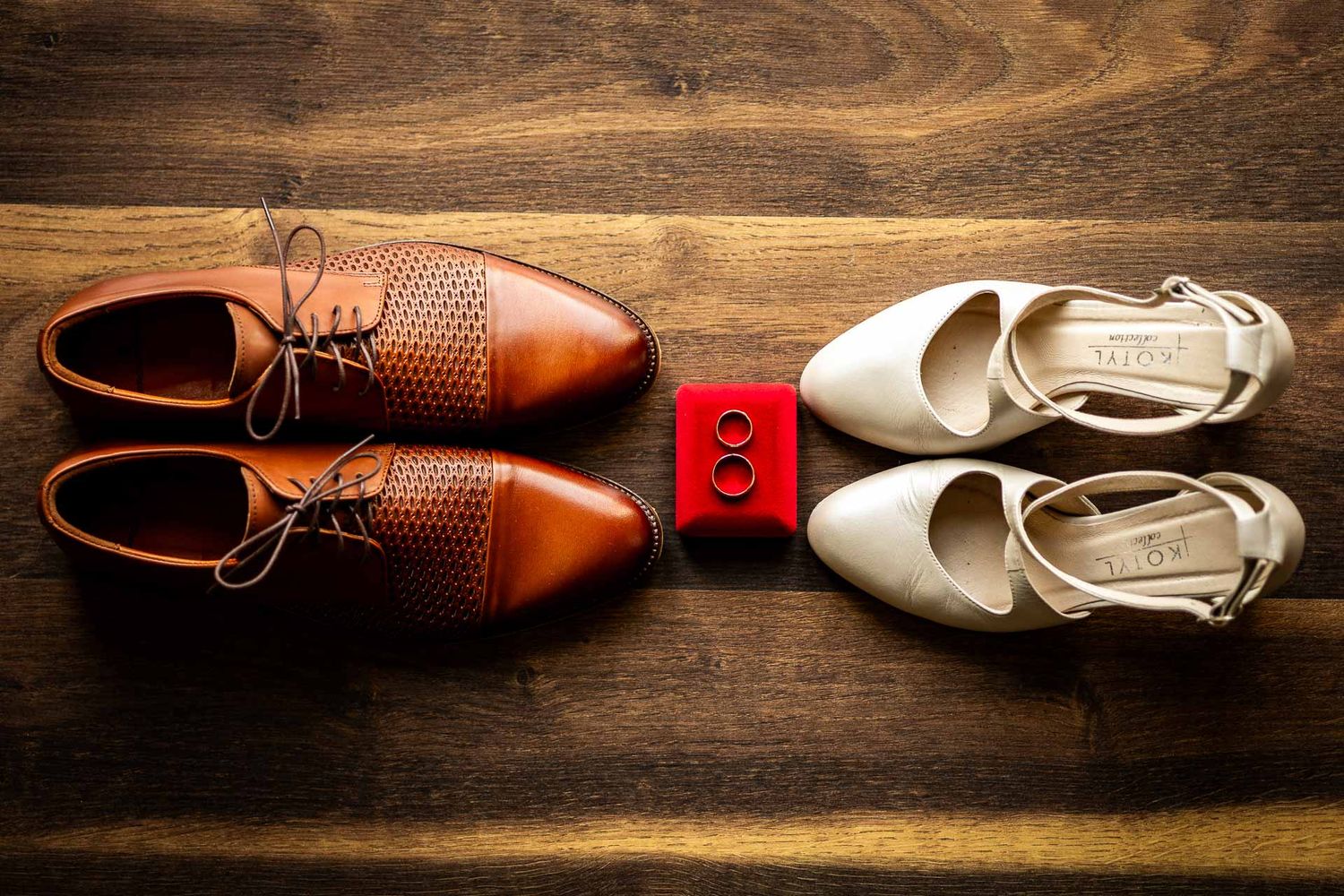 Brown leather dress shoes and white bridal heels arranged on wooden surface for wedding detail shot.