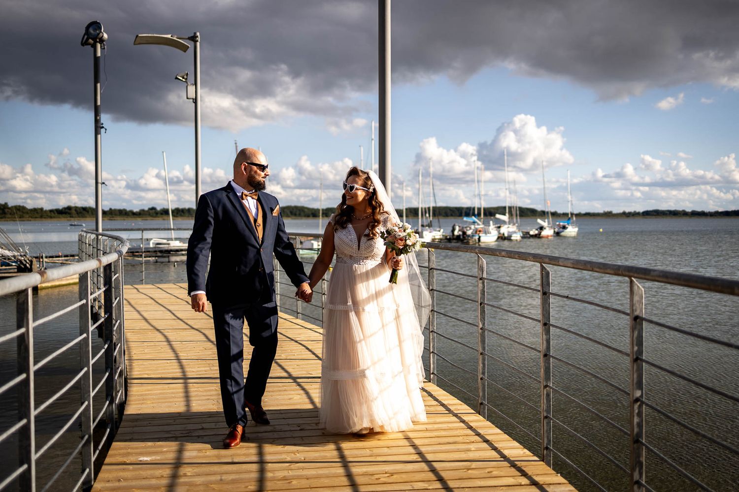Wedding couple walks along a waterfront pier under dramatic cloudy skies.