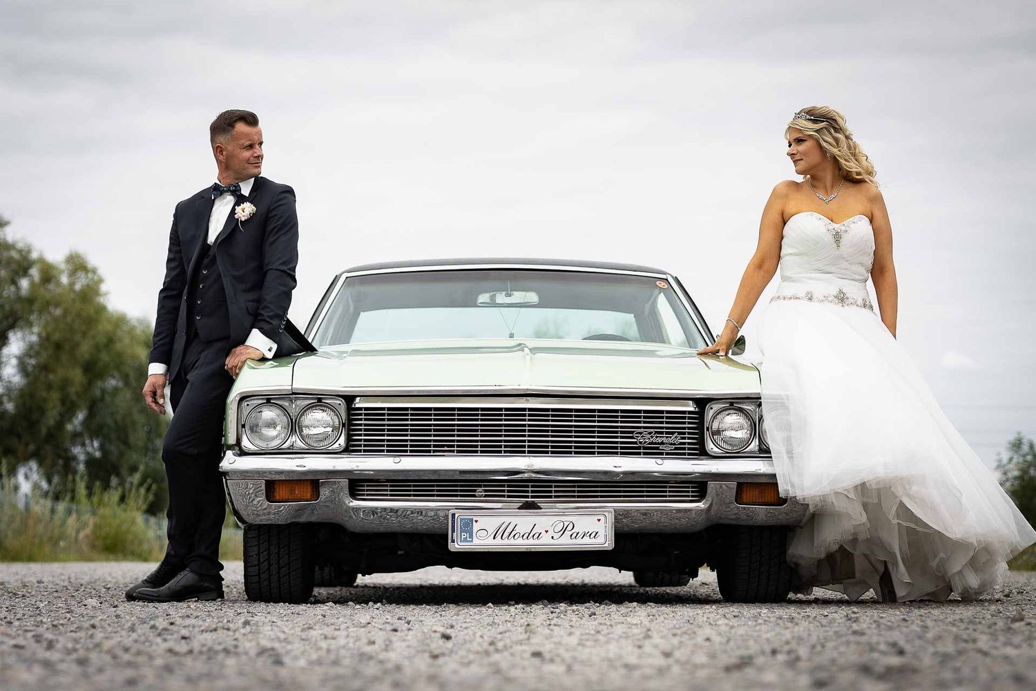 Stylish wedding couple poses with classic Chevrolet car on a rural road.
