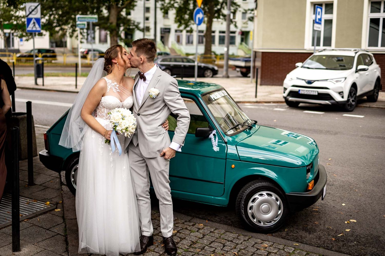 Wedding couple poses romantically next to a vintage turquoise car on a city street.