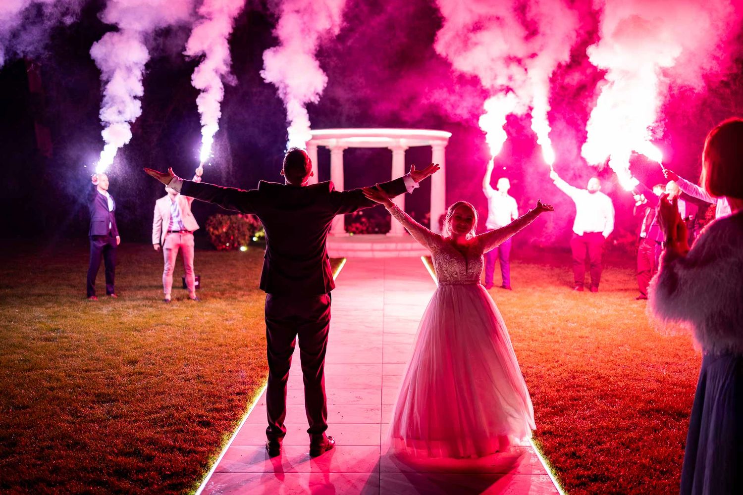 Romantic outdoor wedding scene illuminated by vibrant pink smoke effects.