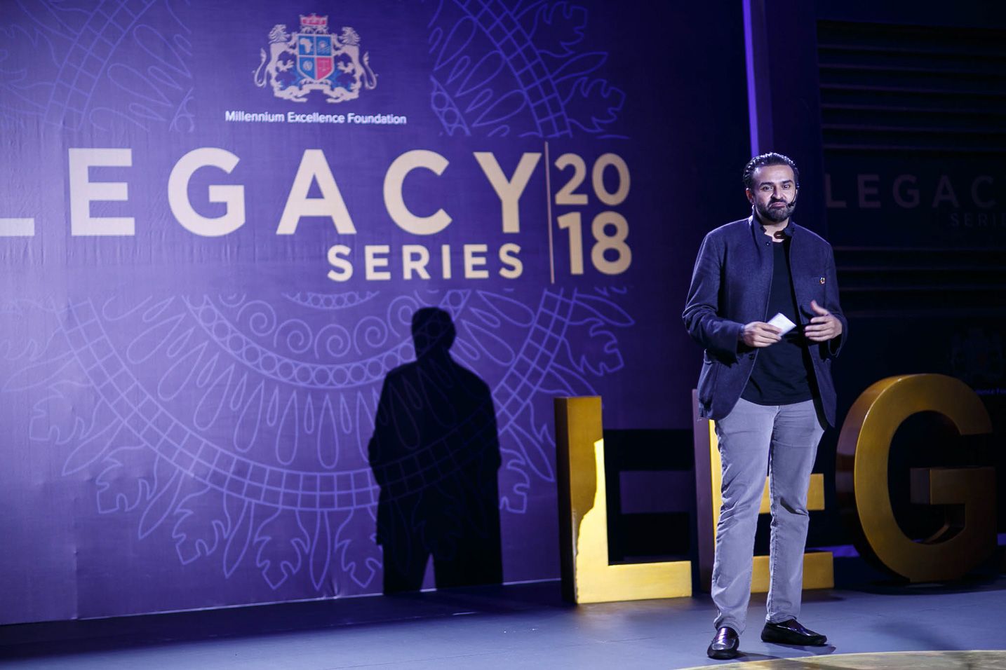 A series of purple-lit stage presentations with silhouettes of speakers against geometric backdrops at a Legacy event.